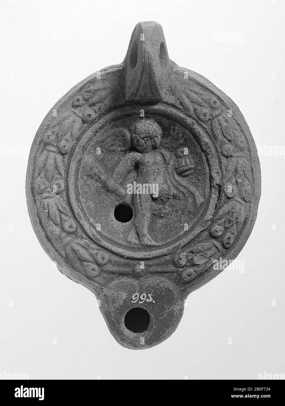 A gray-gray lamp with a round body on a round base. The hollow, round mirror is surrounded with concentric circles and decorated with a relief of a standing Eros with a torch. The small filling hole is located at the bottom left of the figure. The shoulder is decorated with a laurel wreath and berries. The blunt spout is rounded off, has a large burn hole and the incidence is heart-shaped with a relief point and stripe above it. The vertically attached ear is decorated with notches. The underside of the spout is shaded. Brand: two relief points., Oil lamp, earthenware, terracotta, 4.4 x 10.4 x Stock Photo
