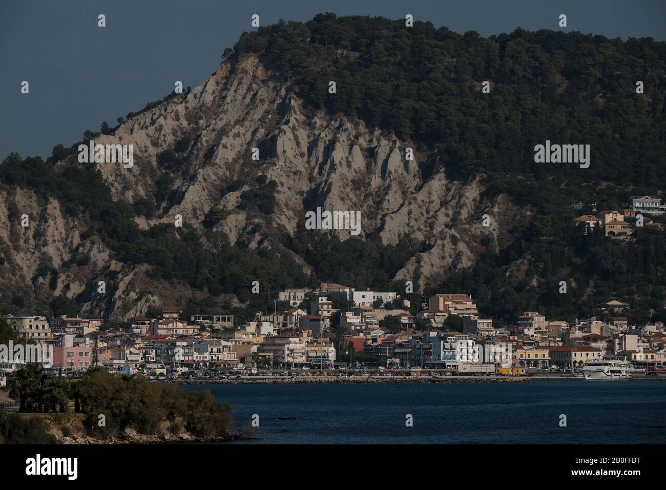 A long lens view of the port of Zakynthos town showing the buildings and harbour overlooked by the dominating limestone cliffs. Stock Photo