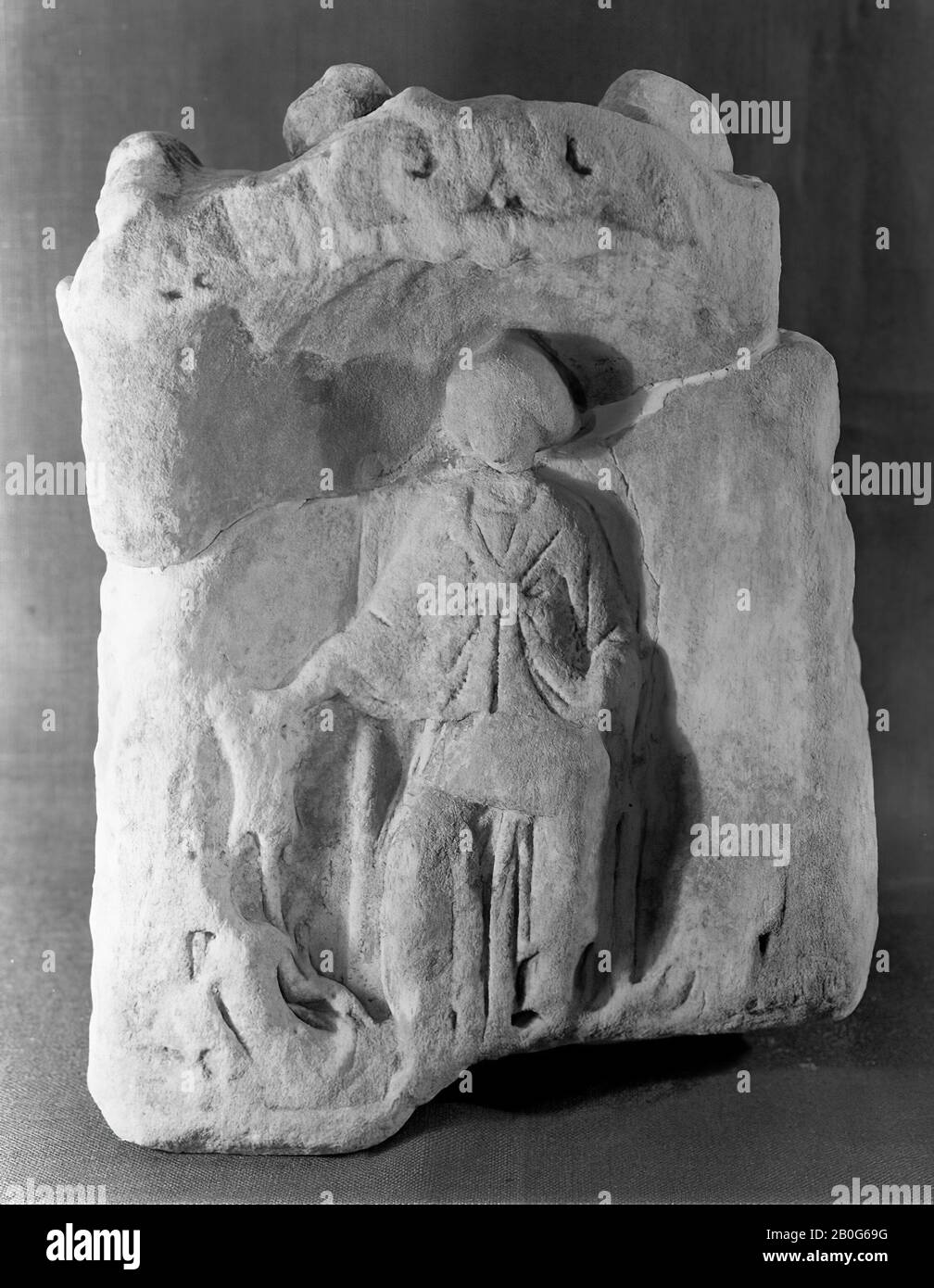 Altar with niche. Sandstone. The lower part with possible inscription is missing. The upper part has been restored from 2 pieces. A corner has broken off at the top right. Nehalennia sitting. The niche is painted blue around the seat with a high back. The shell has remained unpainted. In her right hand she holds a ship's rudder that rests alongside her on the ground. She holds a large object on the left knee. Her clothing is very different than normal, the question is whether there is a shoulder cloak present. On her right side there is a half-seated, half-lying figure on the ground that has a Stock Photo