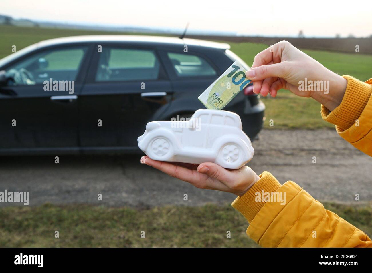 The hand is puting a banknote into the ceramic car shaped money box. The real car is behind. It can be symbol for costs for car´s repairs, investment, Stock Photo