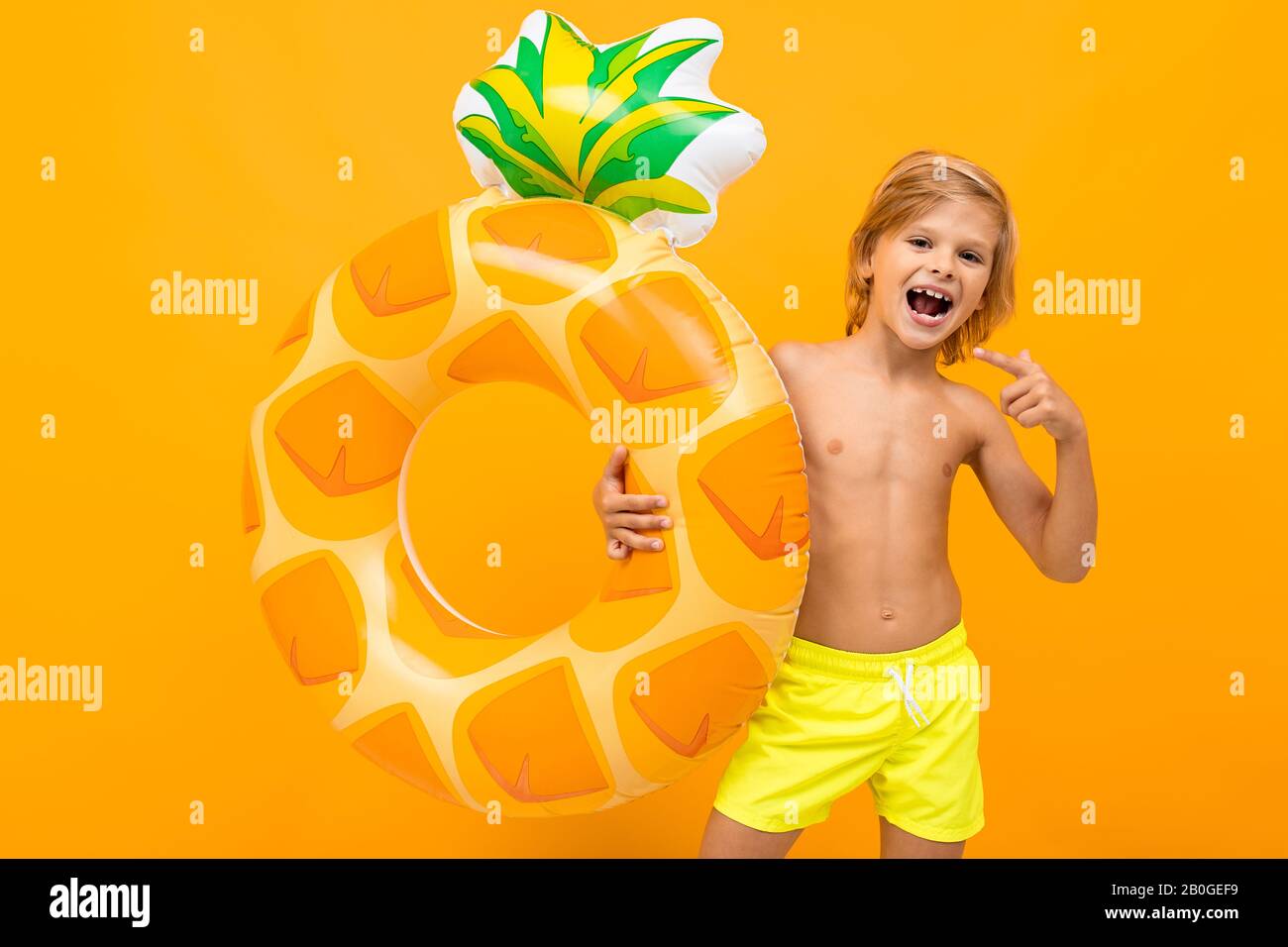Handsome boy in swimming trunks holds a rubber ring, smiles and gesticulates isolated on orange background Stock Photo