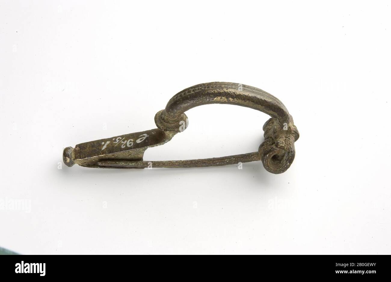 Bronze arch fibula (Haalebos type B), intact, with round brace, which is separated from the long flat foot by an all-round thickening. At the end of the foot a button. Pearl stripe on the bow, fibula, bow fibula, metal, bronze, 5.9 x 2.5 cm, roman 27 BC-41 AD, Netherlands, Gelderland, Neder-Betuwe, Kesteren, Hoge Woerd Stock Photo