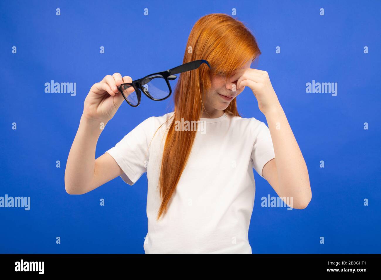 red-haired teenager girl squints while holding glasses in her hand on a blue studio background Stock Photo