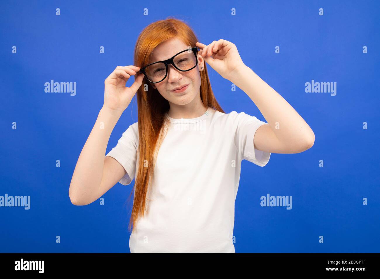 red-haired teenage girl in a white T-shirt squinting holding glasses in his hand on a blue studio background Stock Photo