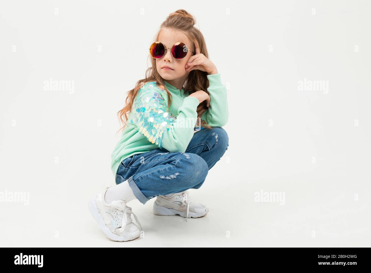 Pretty little girl in blue hoodie and sunglasses sits and smiles isolated on white background