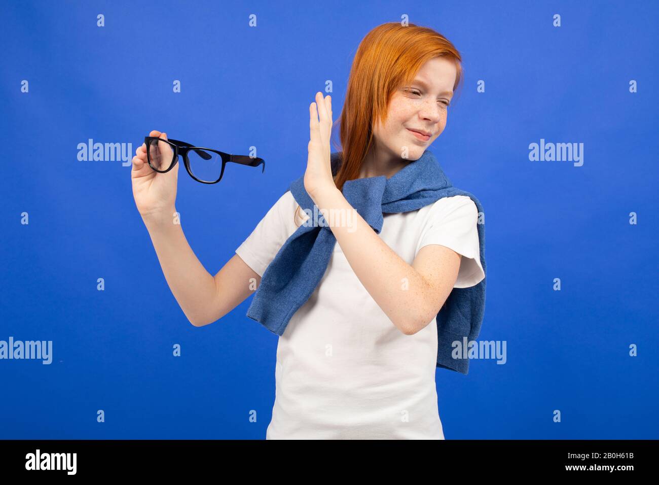 red-haired teenage girl in a white T-shirt throws out glasses on a blue studio background. new vision Stock Photo