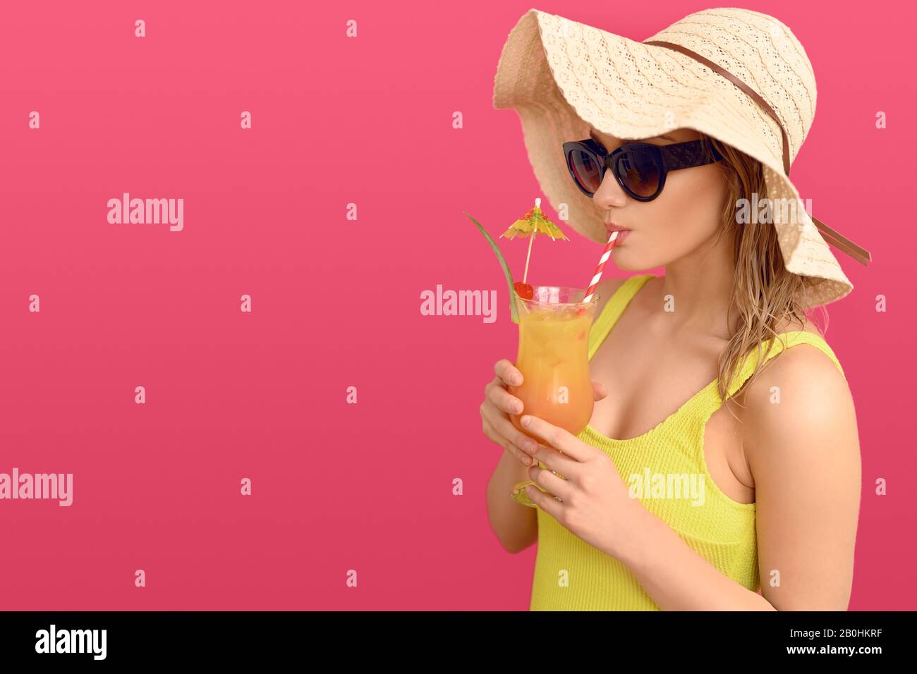 Young woman drinking a tropical cocktail on summer vacation wearing a yellow swimsuit sunglasses and wide brimmed sunhat in a side view over a pink ba Stock Photo