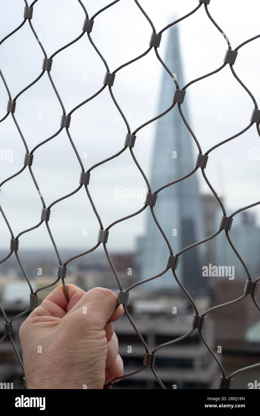 Unusual defocused view of the Shard building London and close up of man's hand gripped wire mesh fence as urban anxiety and uncertainty concept. Stock Photo