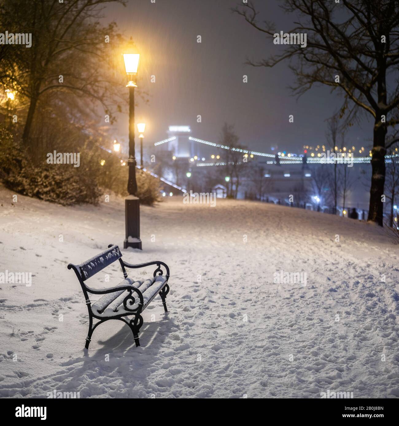 Budapest, Hungary - Bench and lamp post in a snowy park at Buda district with Szechenyi Chain Bridge at background during heavy snowing at winter time Stock Photo