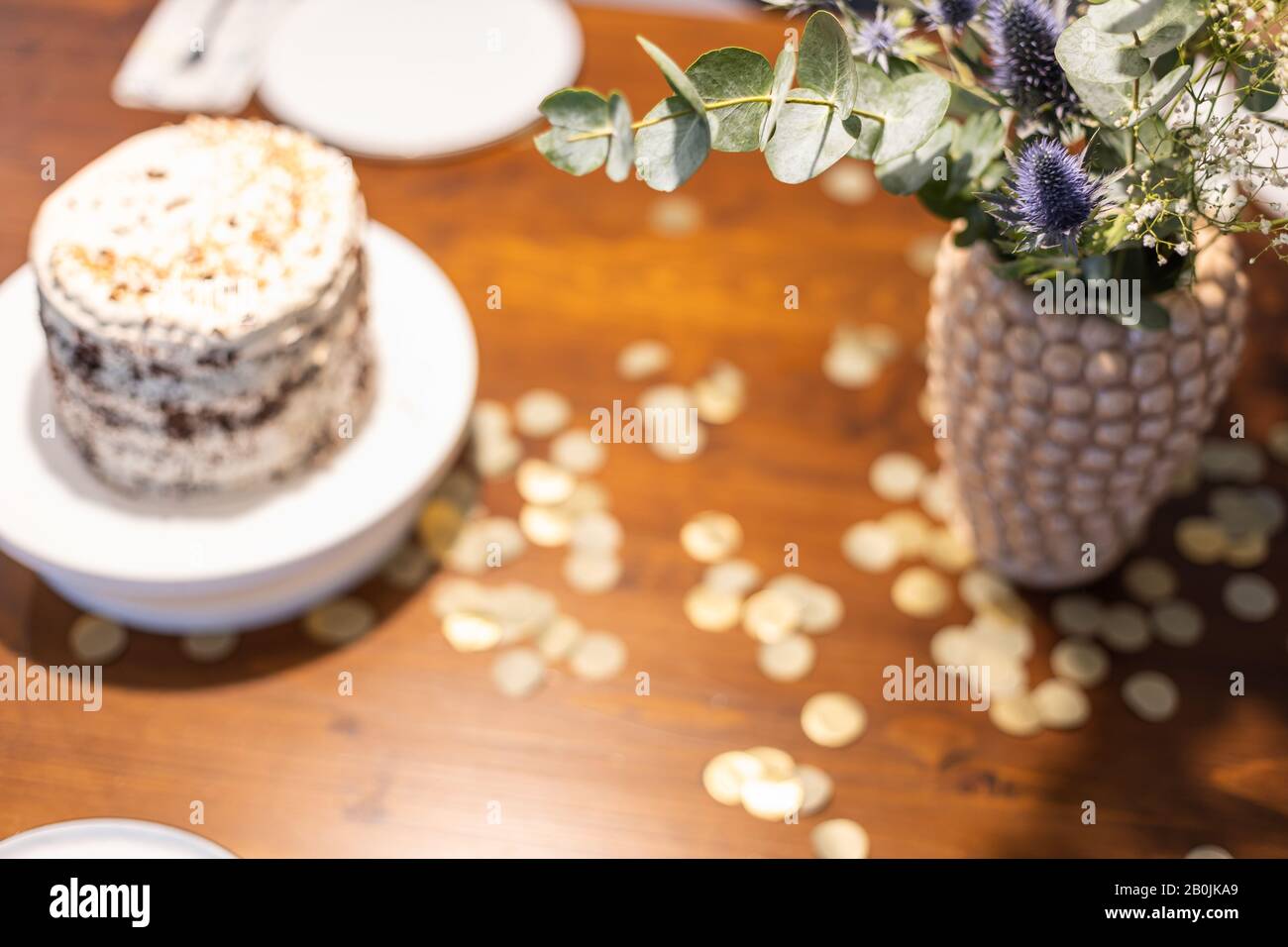 Vase with bouquet of flowers on a table with golden confetti and a naked chocolate cake with sugar pearls. Stock Photo