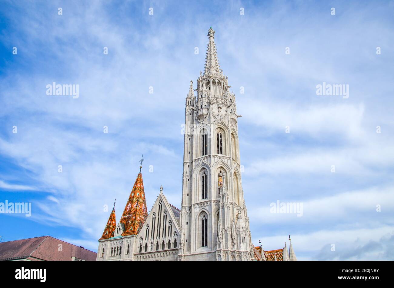 The spire of the famous Matthias Church in Budapest, Hungary. Roman Catholic church built in the Gothic style. Orange colored tile roof. Blue sky and white clouds above. Horizontal photo. Stock Photo