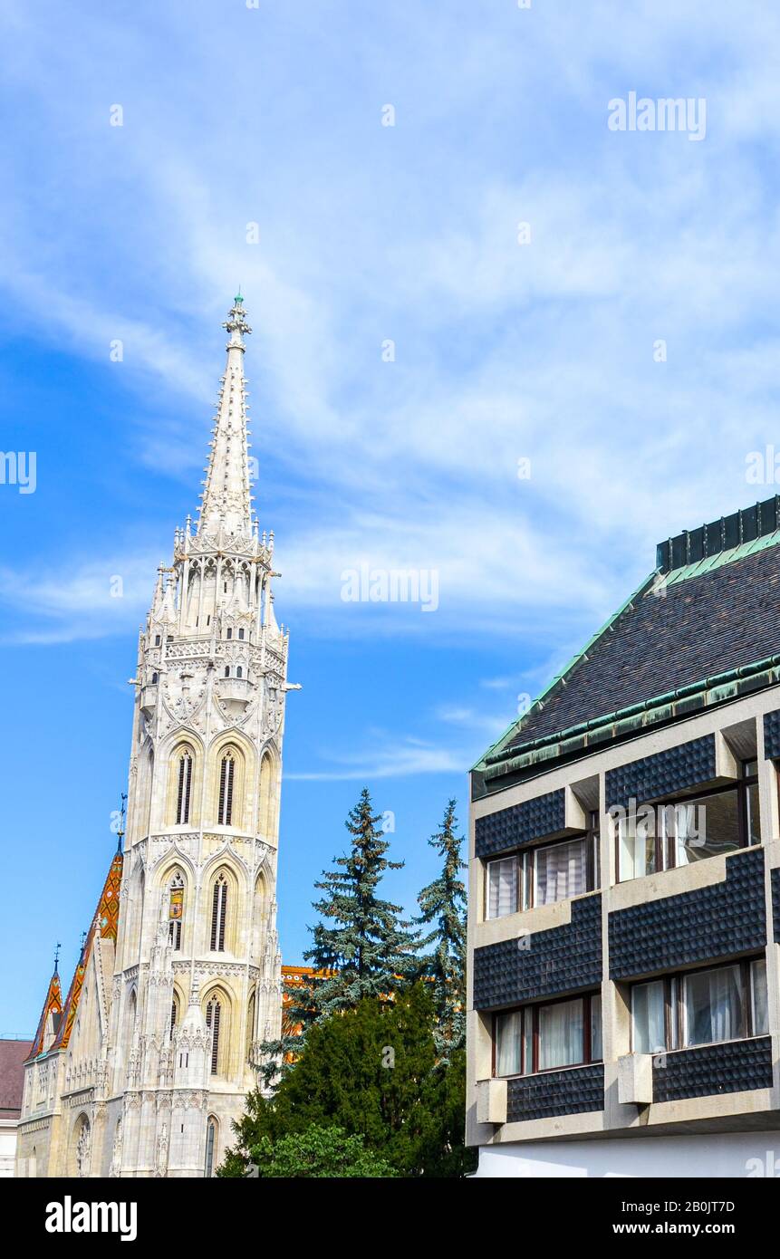 Spire of the Matthias Church in Budapest, Hungary on a vertical photo with adjacent Socialist building. Roman Catholic church built in the Gothic style. Blue sky and white clouds. Eastern Europe. Stock Photo