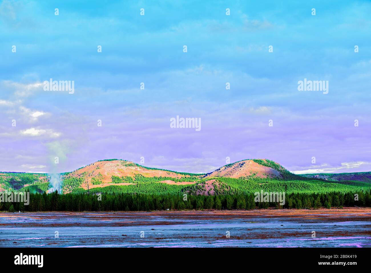 Green forest with smok rising up out of the forest with mountains beyond under purple and blue skies. Stock Photo