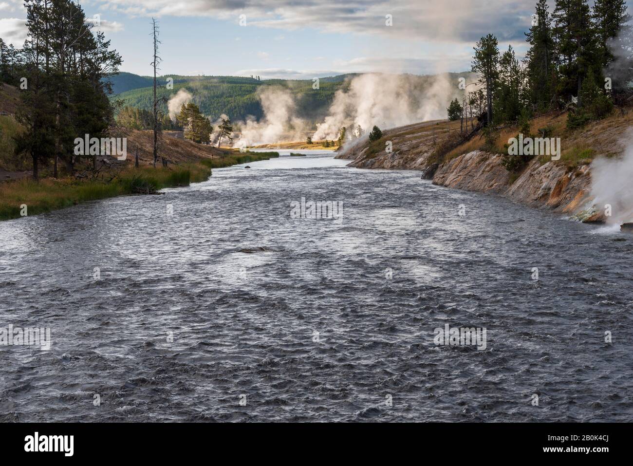 Fast moving river flowing towards geysers venting steam and forested hills. Stock Photo