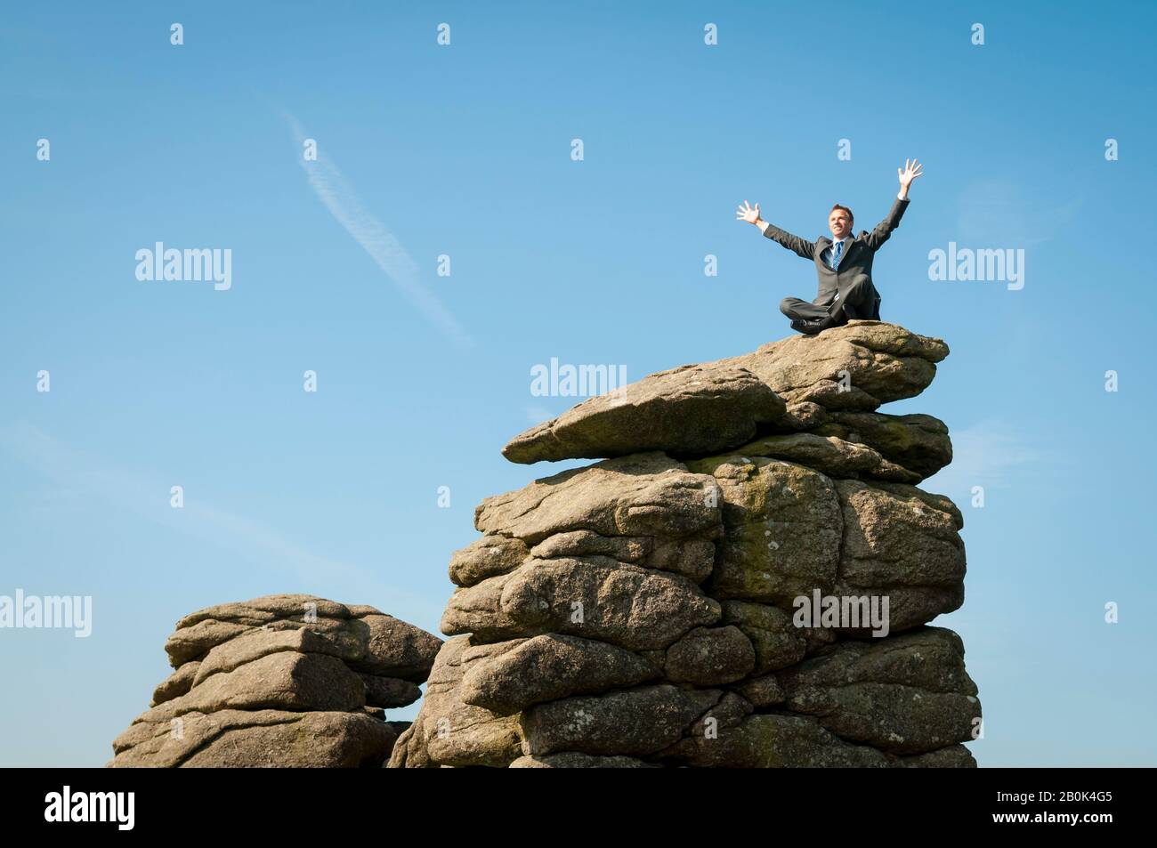Happy businessman celebrating outdoors with arms in the air sitting at the top of a rough rock mountain Stock Photo