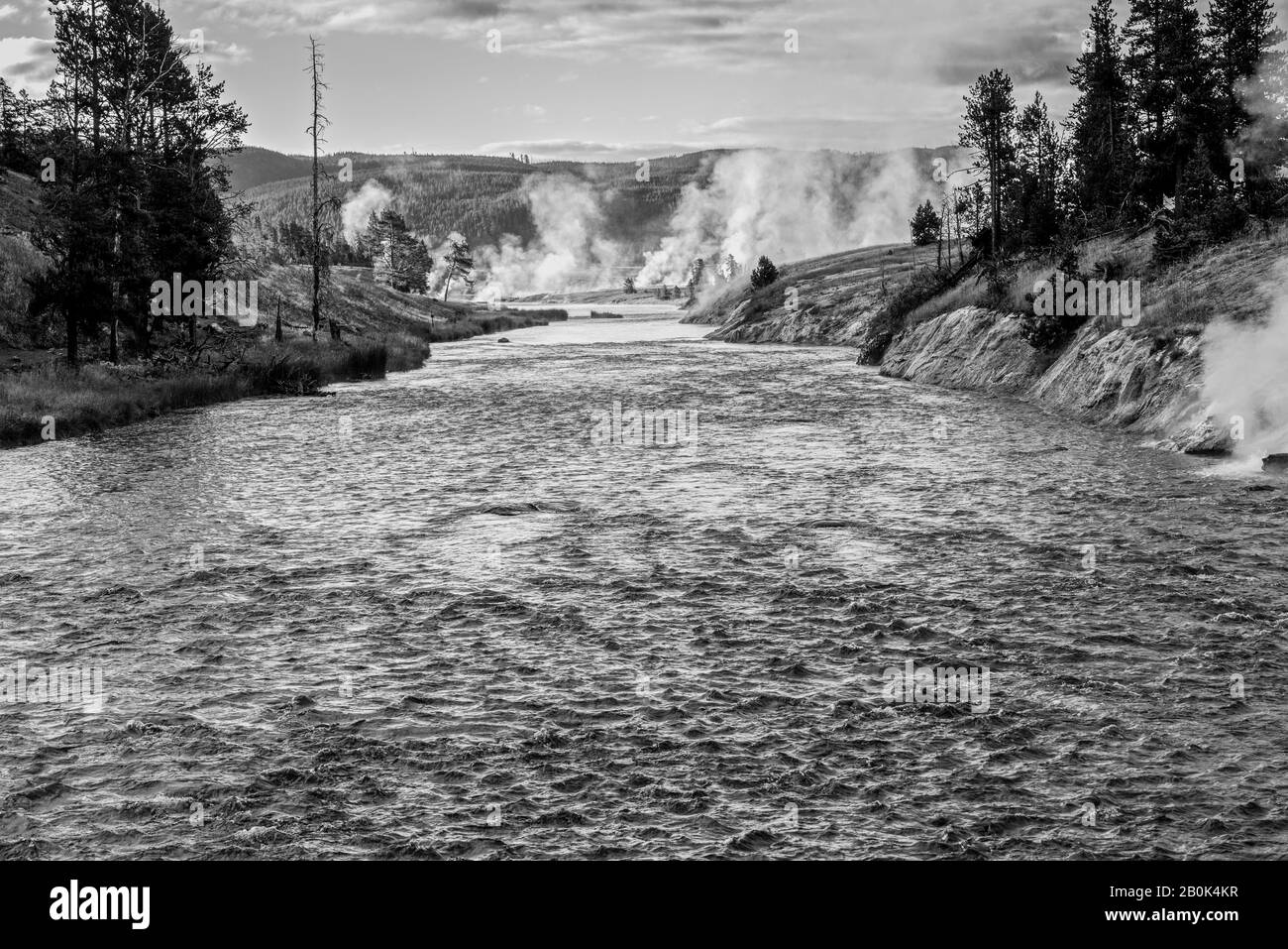 Fast moving river flowing towards geysers venting steam and forested hills. Stock Photo