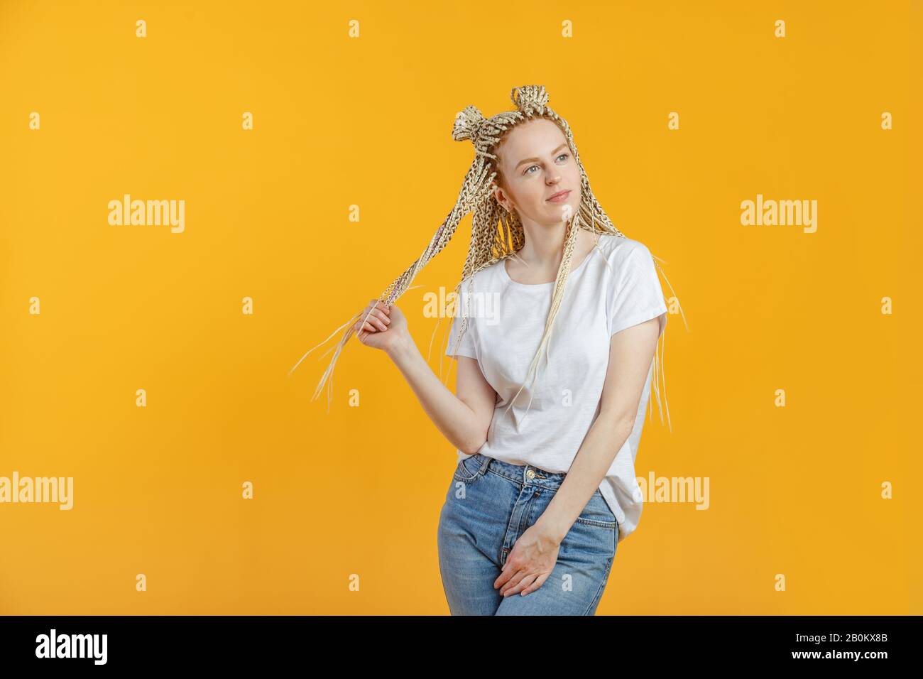 Caucasian young blonde woman dreaming and twisting hair around fingers, corrects isolated yellow background. Stock Photo