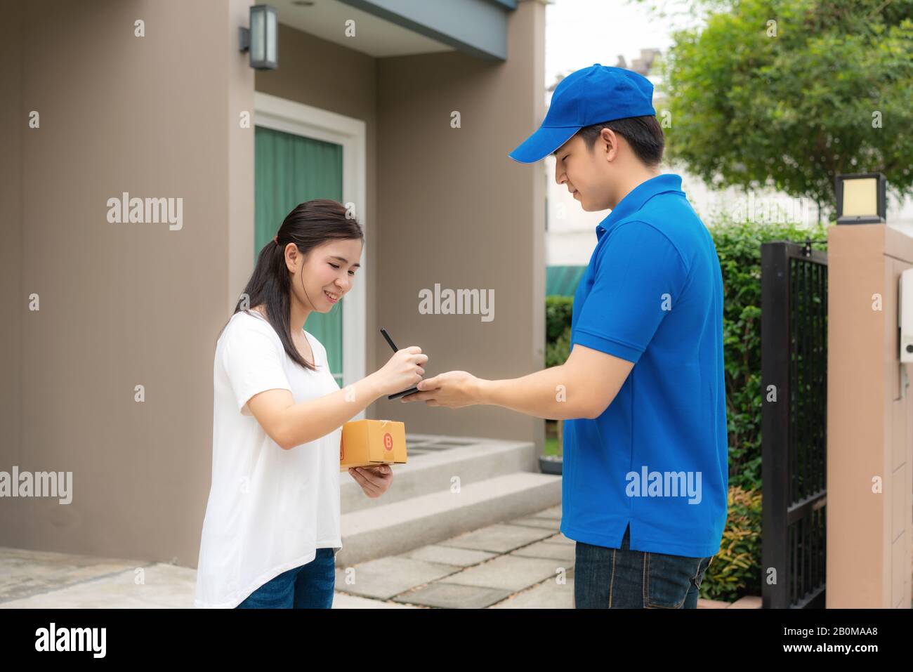 Asian delivery young man in blue uniform smile and holding smartphone in front house and Asian woman sign signature in smartphone to accepting a deliv Stock Photo