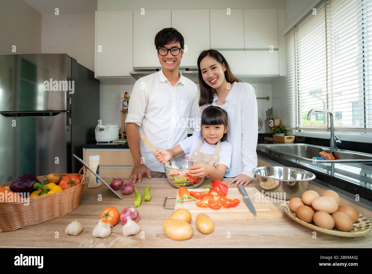 Asian family with father, mother and daughter shredded vegetable salad and look at camera while the family was cooking in the kitchen at home. Family Stock Photo