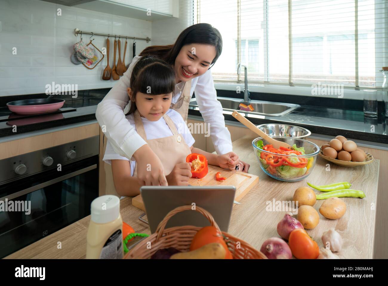 Portrait of beautiful Asian young woman and her daughter cooking salad for lunch using online internet in a digital tablet seach recipe while making f Stock Photo