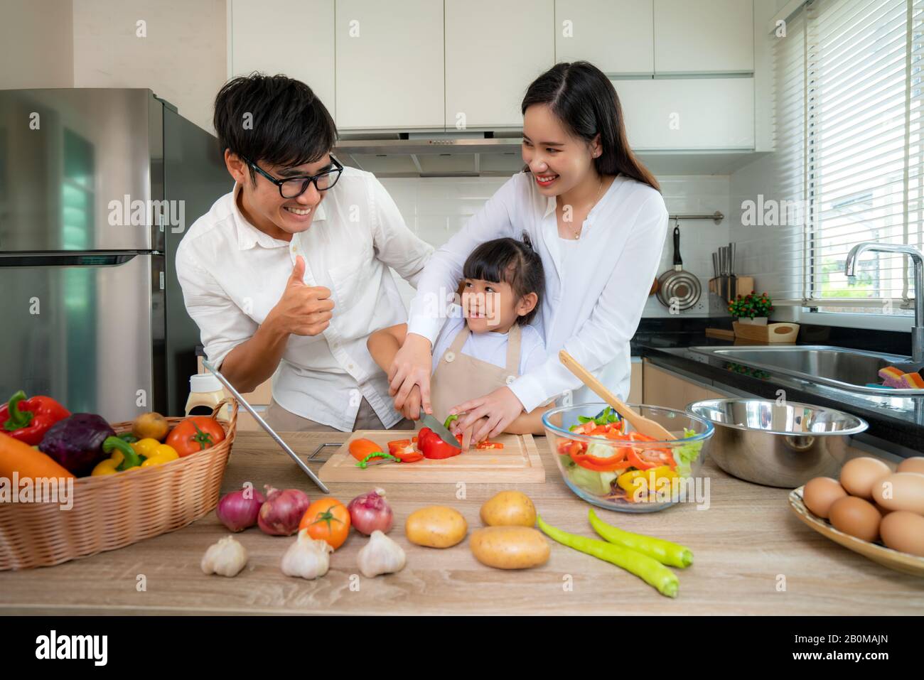 Asian mother teaching her daughter shredded vegetable salad with father thumb up on the side while the family was cooking in the kitchen at home. Fami Stock Photo