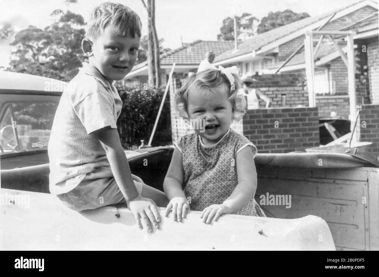 A young Australian boy and girl stand in the back of a Holden FC utility vehicle (Ute) with two male brick layers working in the background in 1965 Stock Photo