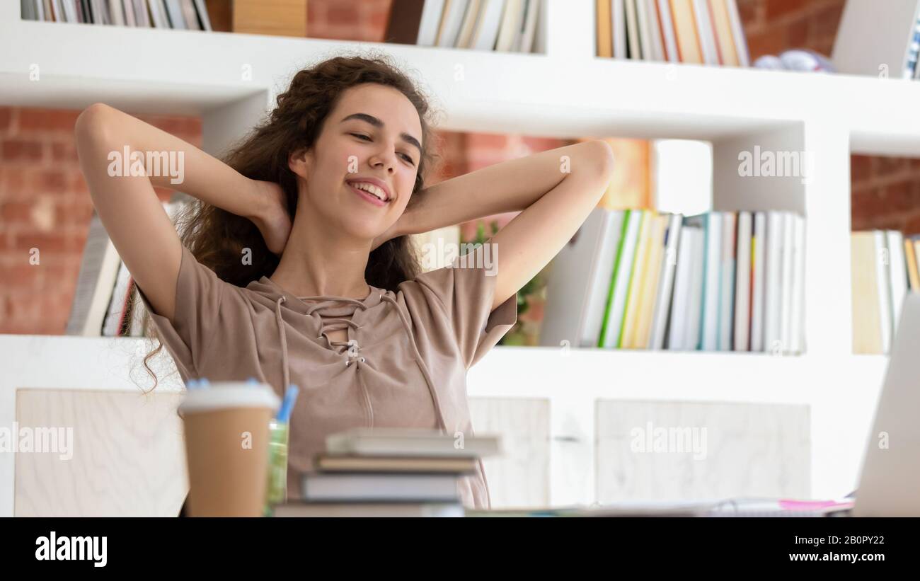 Horizontal photo smiling teenage girl student relaxing during break in library, sitting in comfortable chair, holding hands behind head, woman finishe Stock Photo