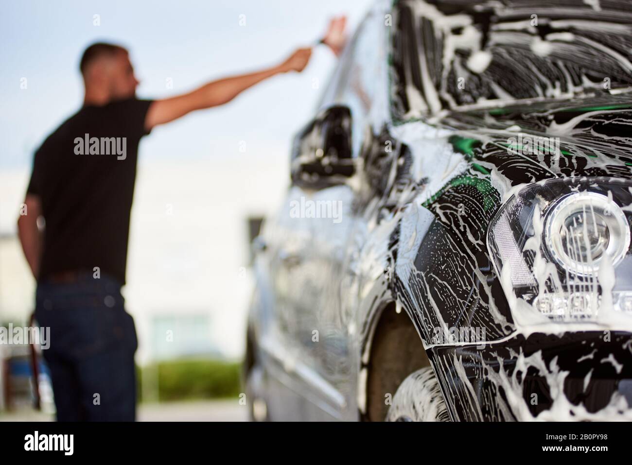 Selective focus on the black car with soap. On the blurred background man is washing his car with a brush and soap outdoors, side view, close-up Stock Photo