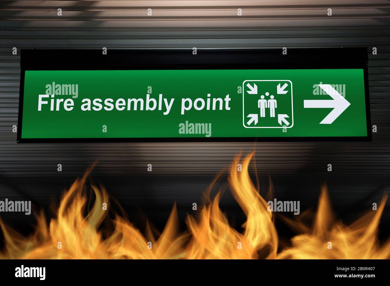 green fire assembly point sign hanging from ceiling with fire flames are burning on the ground below. security based quality awareness concept Stock Photo