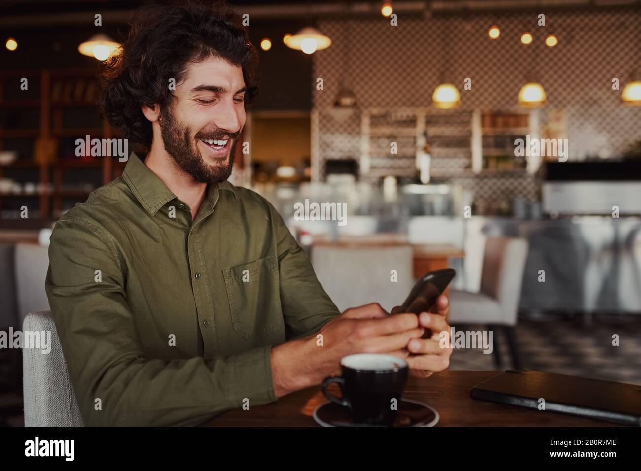 Cheerful handsome young man relaxing in modern cafe using cellphone while drinking coffee Stock Photo