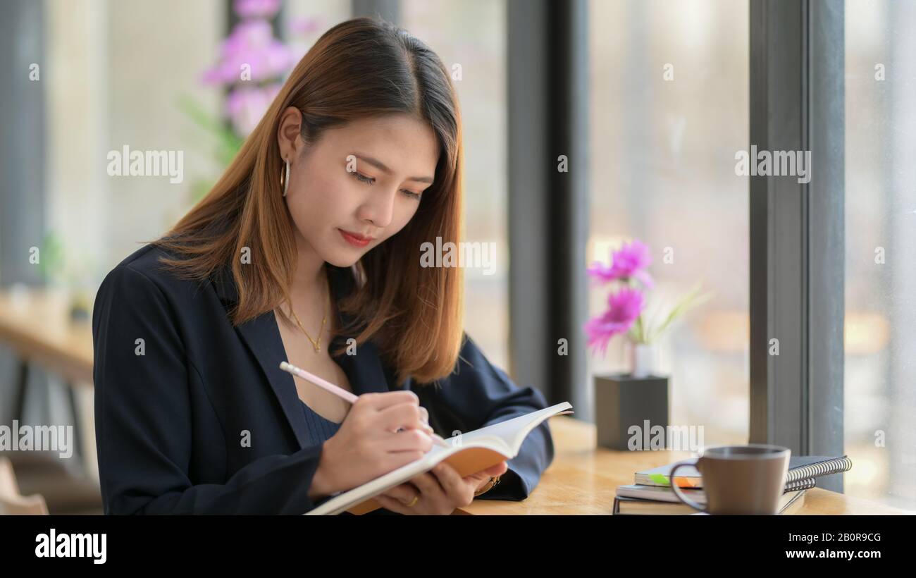 Cropped shot of a woman writing an idea on notebook while sitting on bar stool next window in coffee shop Stock Photo