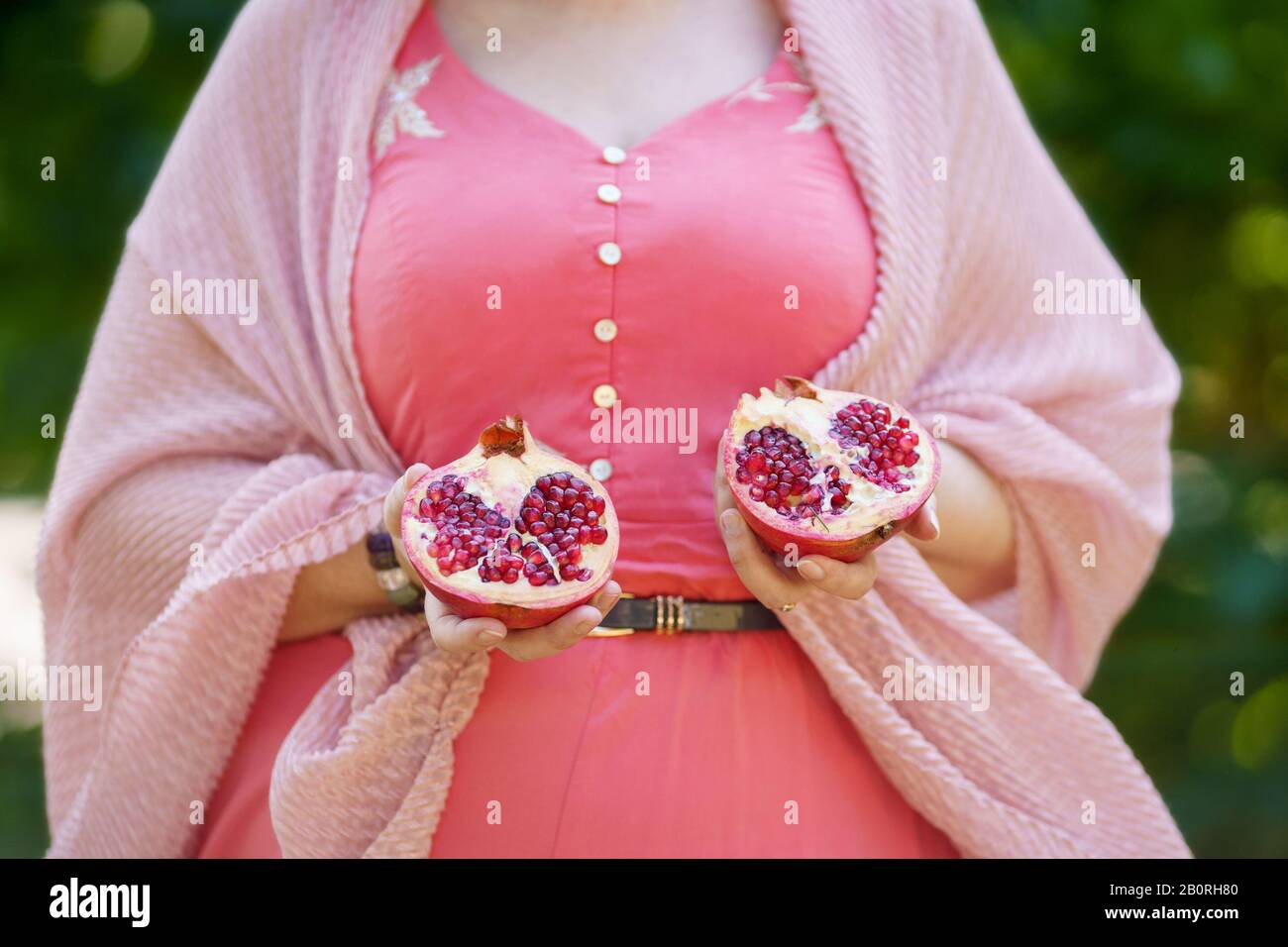 A curvy woman in pink dress and cardigan is holding in her hands two pieces of pomegranate Stock Photo