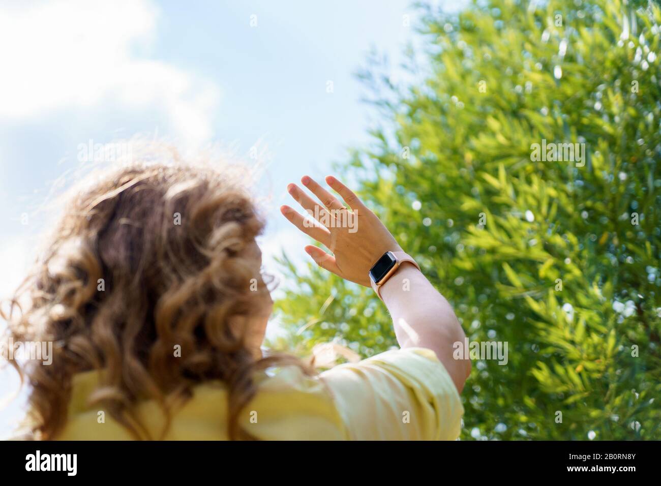 A young woman with red hair in yellow shirt lifted hand with watch up standing her back in summer day against tree in background Stock Photo