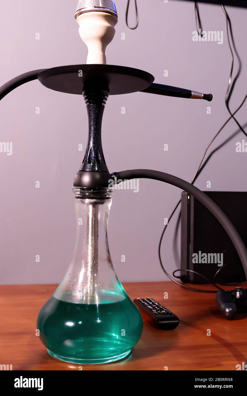 Black matte hookah with clear water with a black hose on the table next to the remote control in the lounge bar on a light background. Hookah cafe lei Stock Photo