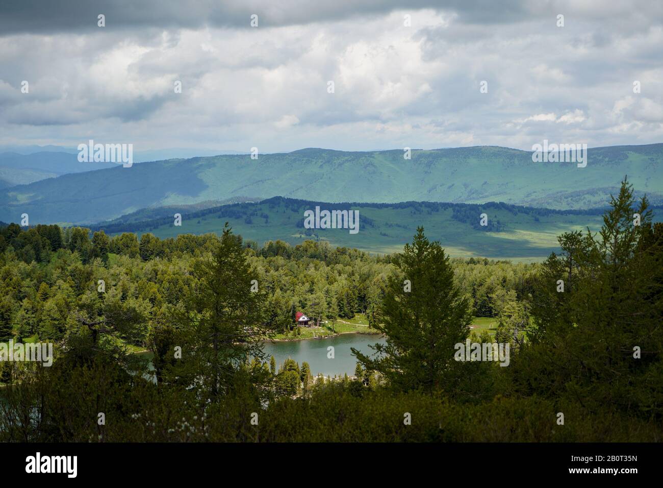 A beautiful mountain lake with small house on the shore under cloudly sky and mountains growned by the forest on the background Stock Photo