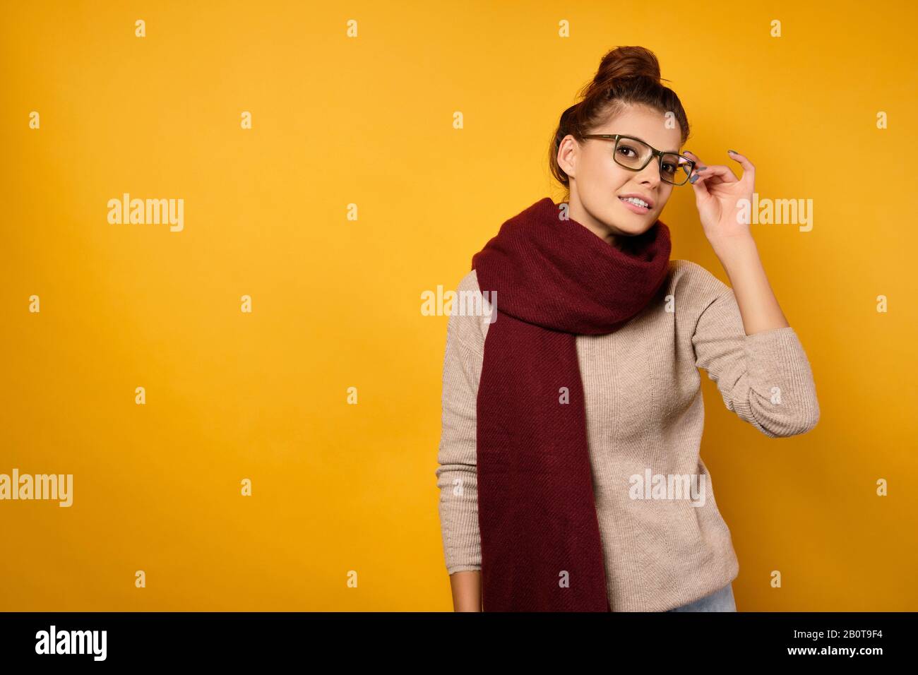 A beautiful brunette with a bun on head and glasses stands in a sweater and a burgundy scarf, looks at the camera with head bowed Stock Photo