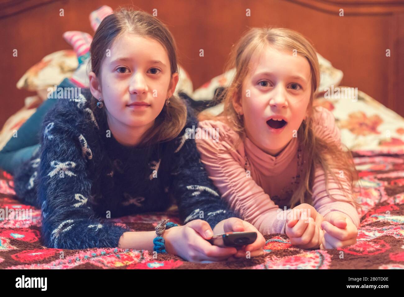 Two happy girls watching tv or movie. A horizontal photo of two young pretty smiling sisters lying on bed and watching their favourite serials. Stock Photo