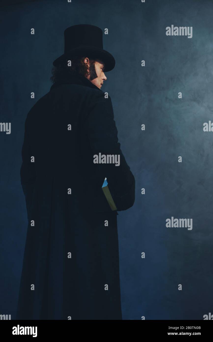 Edwardian man in long black coat and hat holding book standing towards