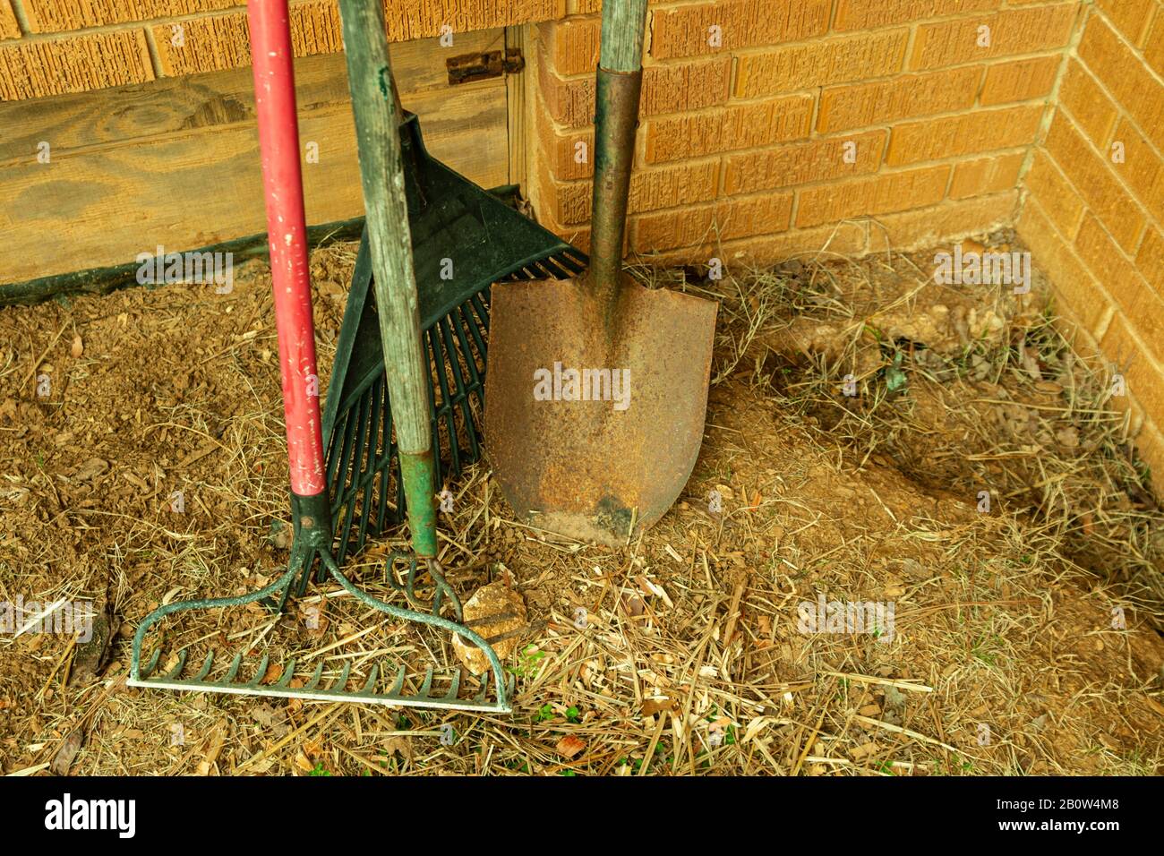 A garden rake, leaf rake, hoe and rusty shovel leaning against a brick