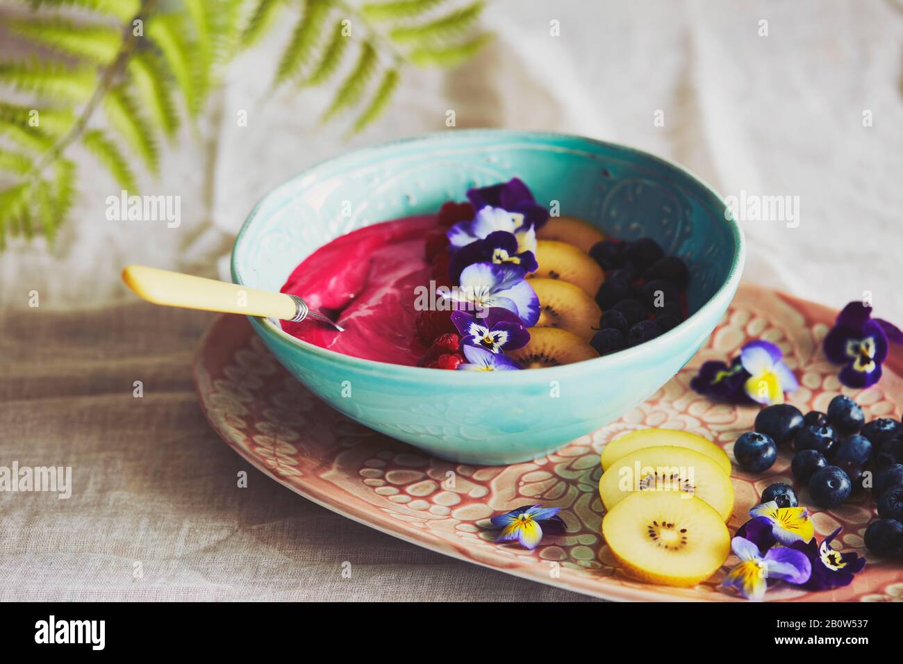 High angle close up of bowl with pink fruit puree with blueberries and slices of yellow kiwis, decorated with pansy petals. Stock Photo