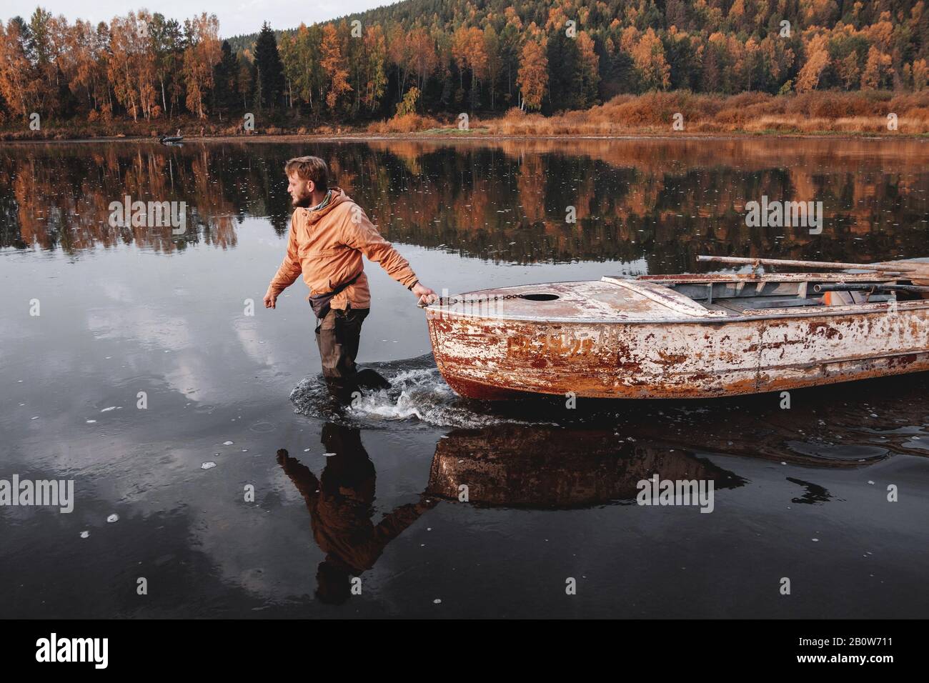 Man dragging old rusty boat in middle of lake in autumn, Russia Stock Photo