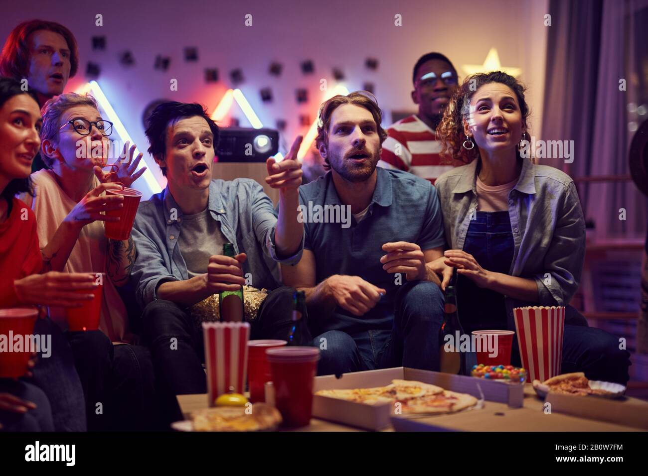 Group of young people sitting on sofa eating pizza and popcorn and watching a movie together during home party Stock Photo