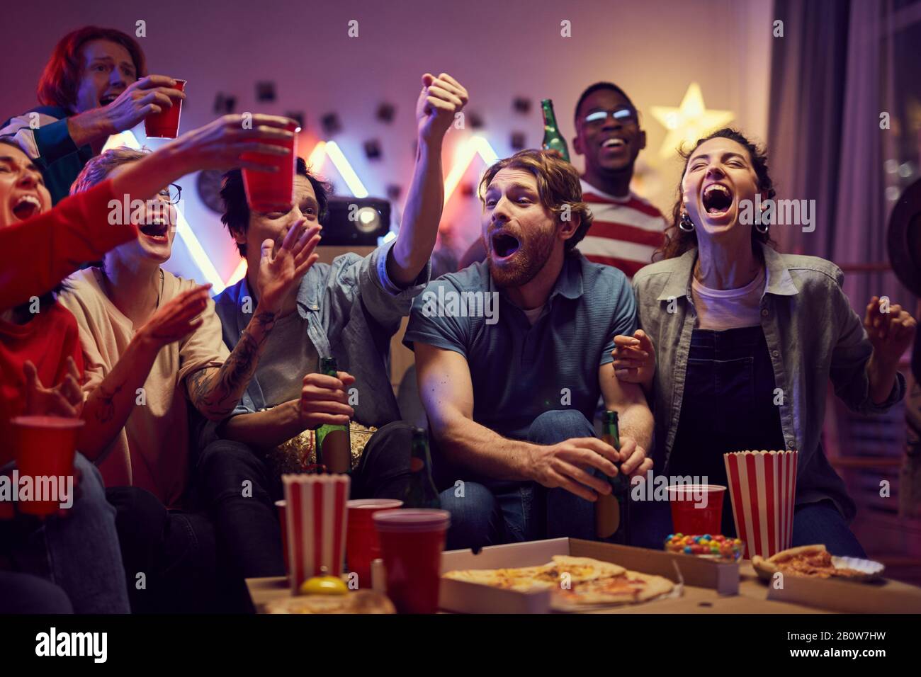 Group of young fans sitting on sofa drinking beer and eating pizza while watching football match on TV at home Stock Photo