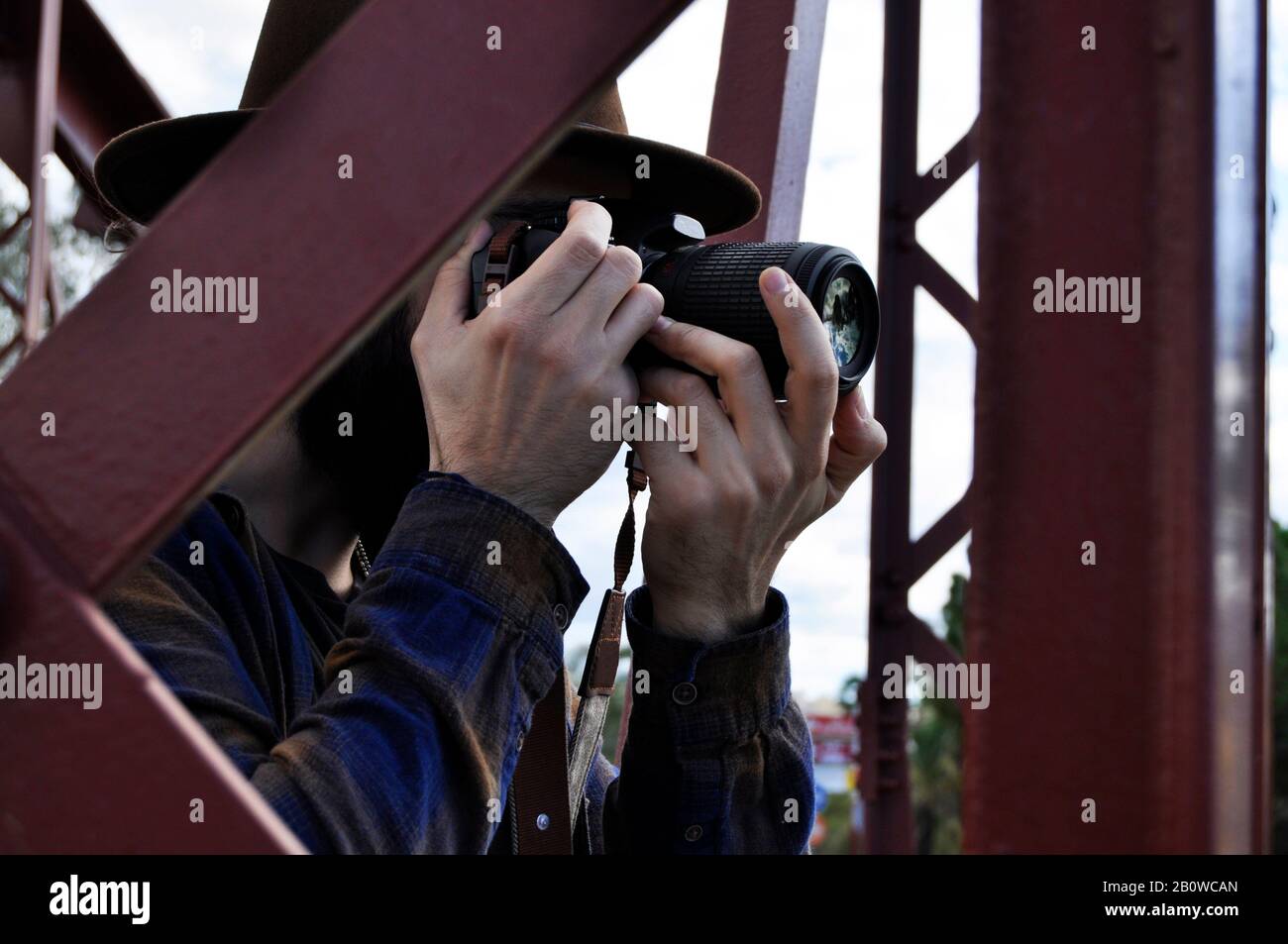 Man photographer taking a photograph through a big red metal building bridge wearing a brown cowboy hat and a squares shirt, modern people Stock Photo