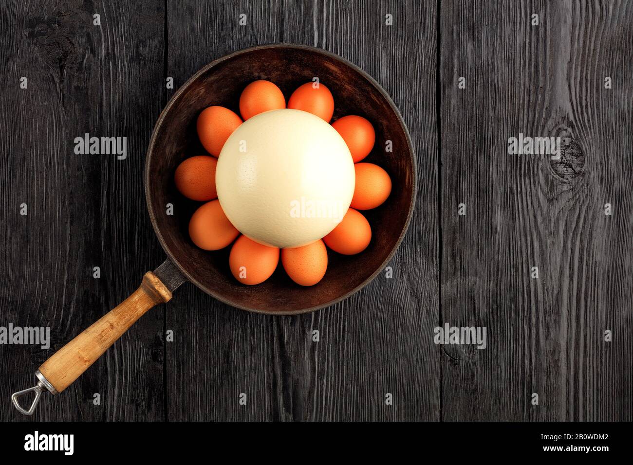 Ostrich egg surrounded by chicken eggs in an old cast-iron skillet, which is standing on an old black wooden surface, top view, copy space. Stock Photo