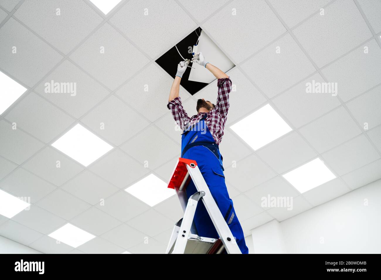 Full Length Portrait Of Electrician On Stepladder Installs Lighting To The Ceiling In Office Stock Photo