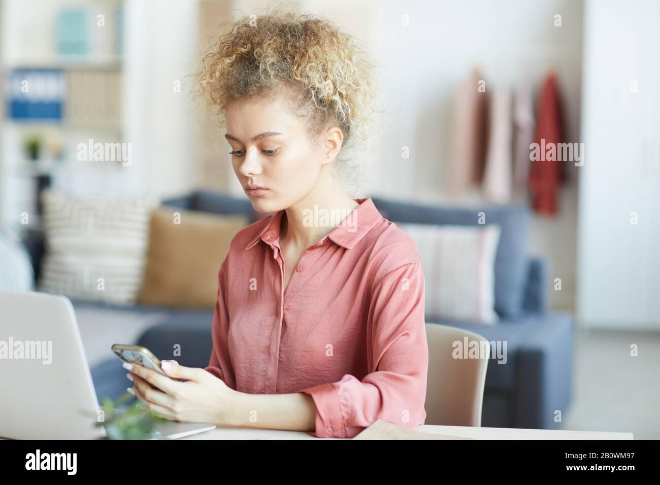 Young woman with curly blonde hair sitting at the table and using her mobile phone at home Stock Photo
