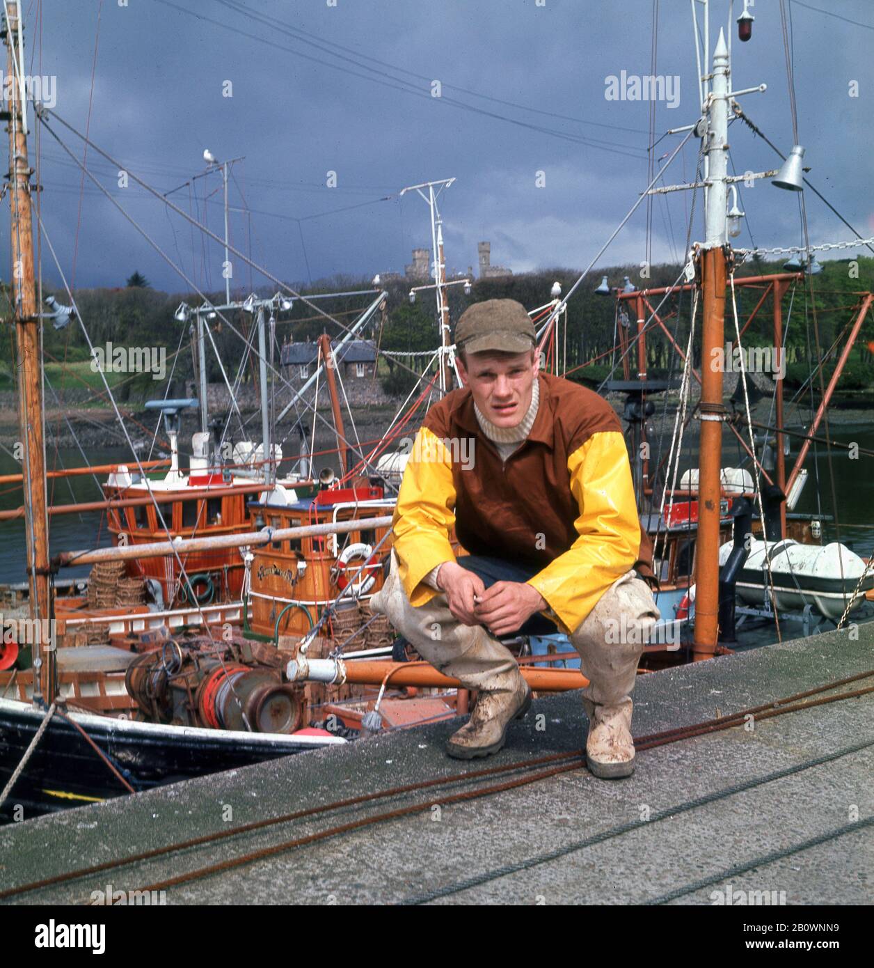 1960s, historical, a young Scottish fisherman in his waterproof fishing clothes and cap, kneeling down for a photo dockside at the port of Stornoway, Isle of Lewis, Scottish HIghland, Scotland, UK. Stock Photo