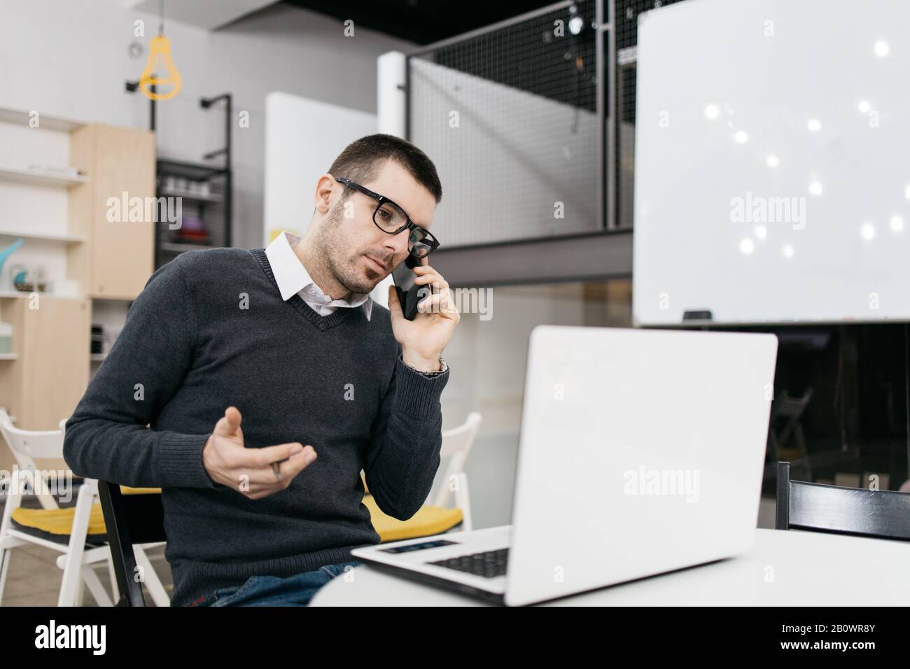 Man with glasses using phone and making gestures. Office worker using laptop while talking on phone and doing hand gestures. Stock Photo