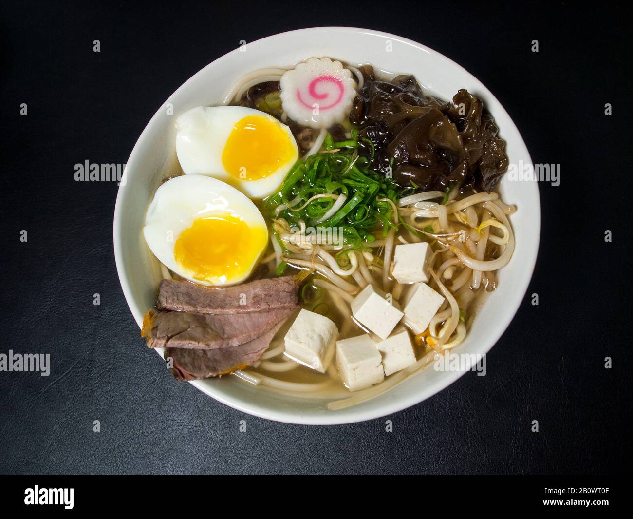 Ramen: Asian noodle soup with beef, eggs, fungi, vegetables, tofu, seaweed and naruto in a bowl. Black background. Top view. Stock Photo